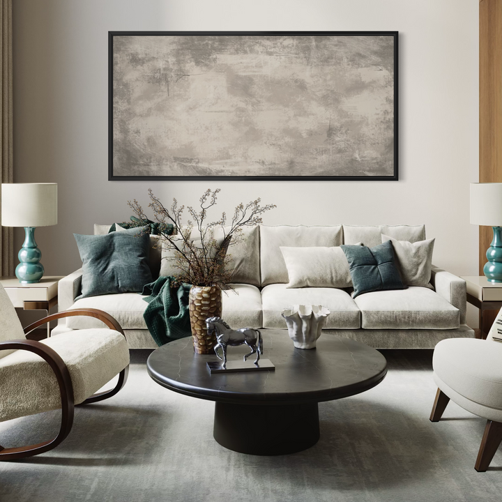 A large framed abstract canvas print with textured gray, beige, and brown tones hangs on a light beige wall above a modern cream-colored sofa in a styled living room.