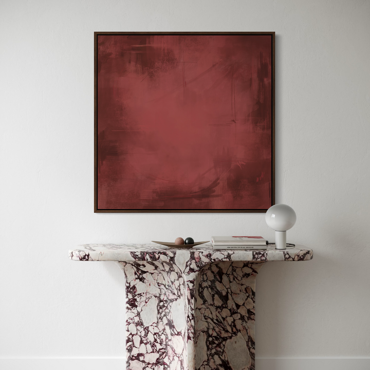 A square framed abstract painting in deep burgundy red hangs on a white wall above a white and burgundy marble console table with a white globe lamp.