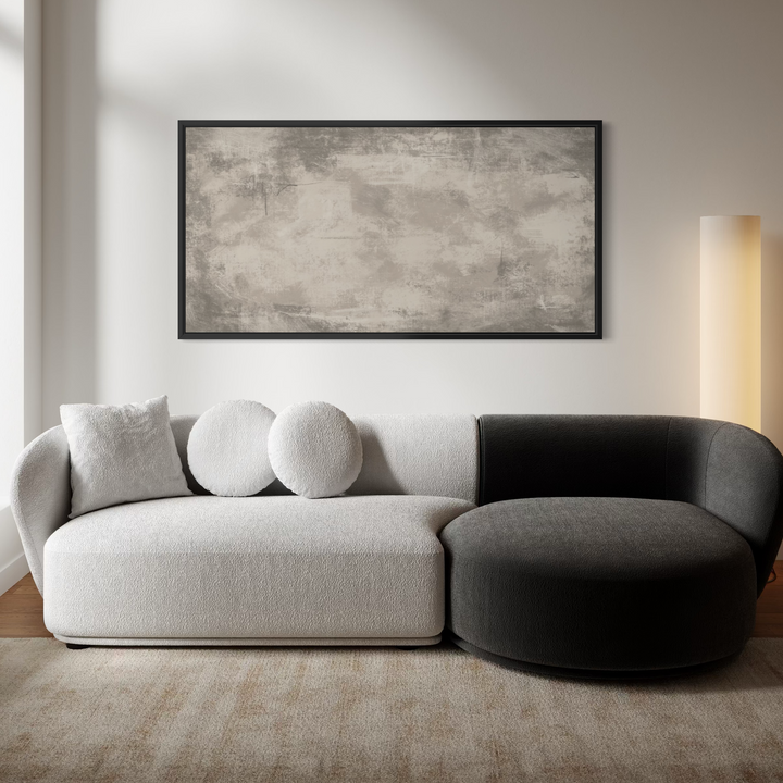 A modern sectional sofa in a minimalist living room, featuring a light beige textured fabric on the left and a dark charcoal gray on the right, with three round throw pillows. A large framed abstract canvas print hangs on the wall above the sofa.
