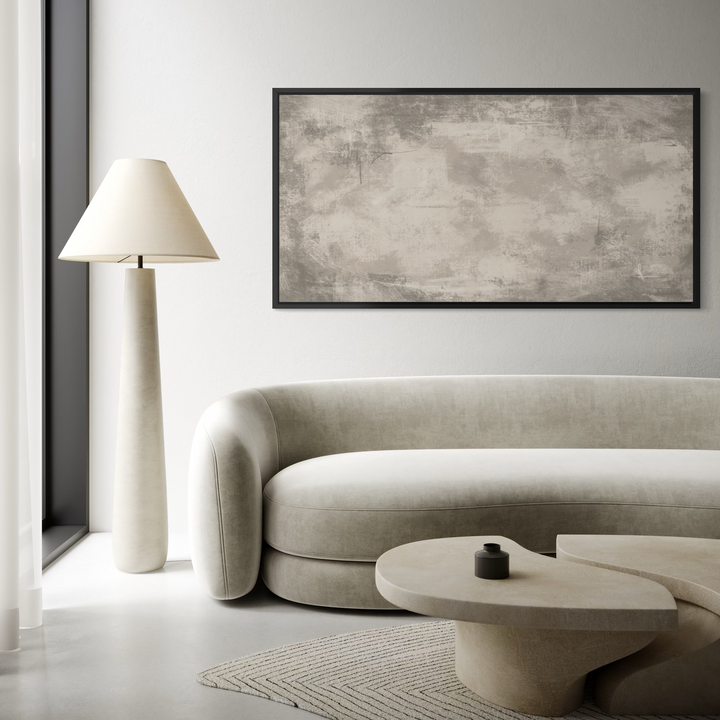 A modern living room scene featuring a large abstract framed wall art print with textured beige and gray tones, hung above a curved light beige sofa. The room is styled with a tall white floor lamp, a low organic-shaped stone coffee table, and a textured rug.