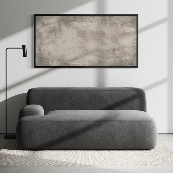 A modern gray upholstered daybed or chaise lounge in a minimalist living room setting, positioned below a large framed abstract canvas print. A black floor lamp stands to the left, and strong diagonal sunlight casts shadows across the sofa and wall.