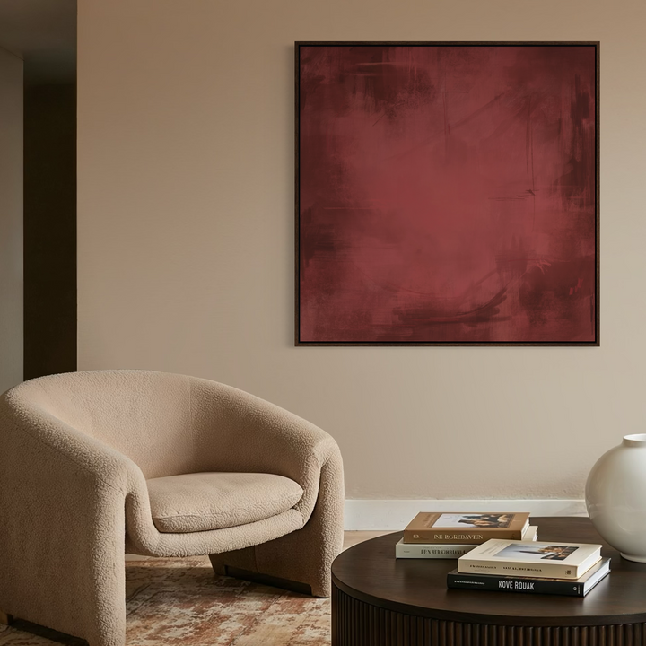 A framed abstract square painting in deep burgundy red hangs on a beige wall in a styled living room setting with a cream boucle armchair, a dark wood coffee table with books and a white vase, and a patterned rug.