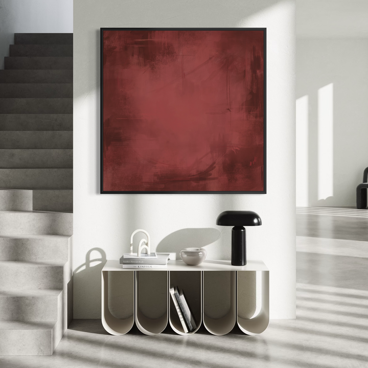 A large square abstract wall art piece in deep red hangs above a sculptural white sideboard in a modern interior with gray stairs, a black table lamp, and soft window shadows.