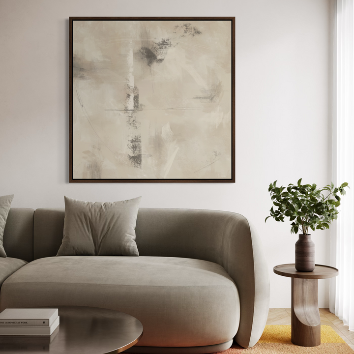 A large square framed abstract wall art print with soft beige, cream, and gray brushstrokes hangs above a modern gray sofa in a bright, minimalist living room. A small wooden side table with a potted green plant sits to the right.