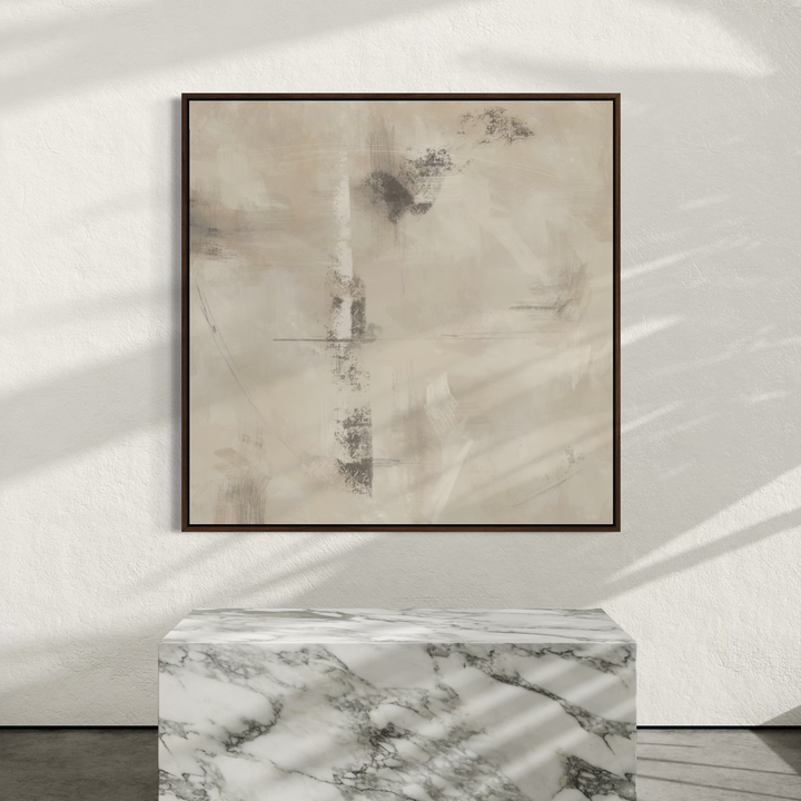 A square framed abstract wall art print with soft beige, gray, and brown brushstrokes, displayed on a white wall above a white-and-gray marble pedestal. Sunlight casts leaf-like shadows across the wall and pedestal.